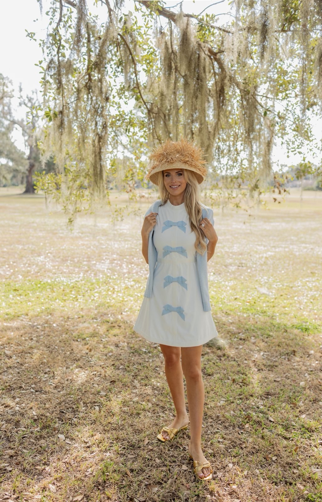 Woman in a striped dress and hat standing in a park with trees and grass.