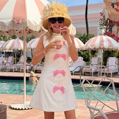 Woman in a white dress with pink bows standing by a poolside with palm trees and striped umbrellas.
