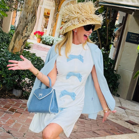 Woman in a light blue dress with dolphin pattern, straw hat, and sunglasses standing outdoors.