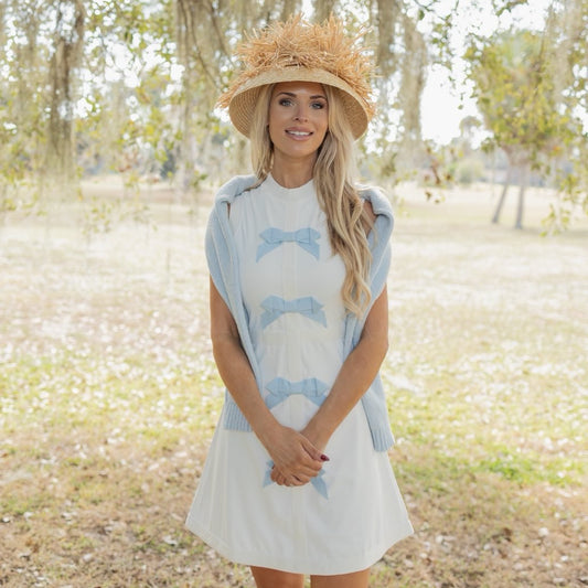 Woman wearing a light blue dress with bow patterns and a straw hat in a park setting.