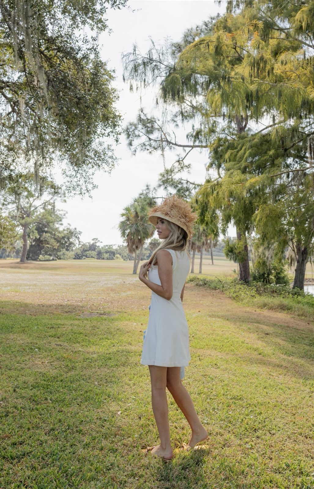 Woman in a white dress and straw hat standing in a park with trees and grass.