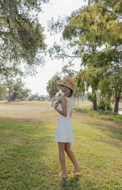 Woman in a white dress and straw hat standing in a park with trees and grass.