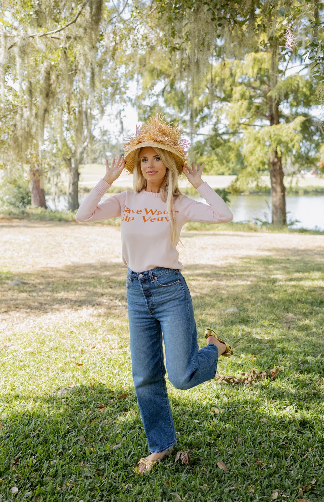 Woman in a park wearing a straw hat, light pink shirt, and blue jeans.
