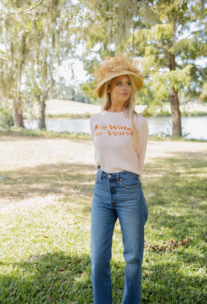 Woman wearing a white t-shirt with text and blue jeans, standing outdoors with trees and water in the background.