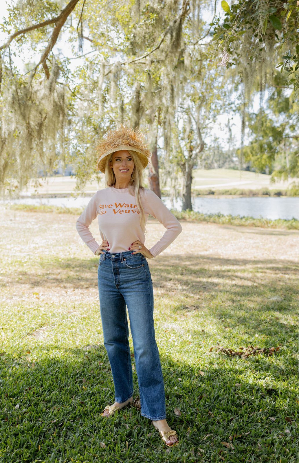 Woman wearing a white long-sleeve shirt with text and blue jeans, standing in a park with trees and a pond in the background.