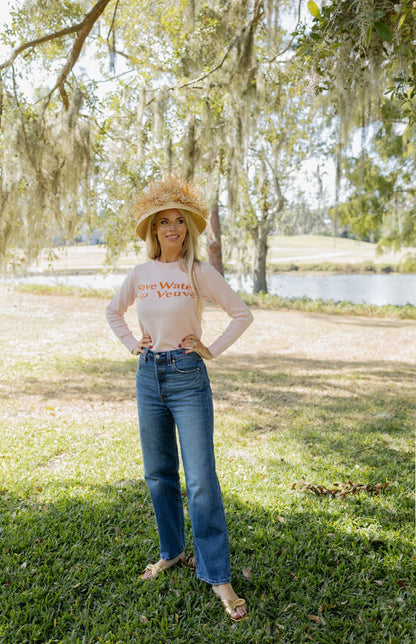 Woman wearing a white long-sleeve shirt with text and blue jeans, standing in a park with trees and a pond in the background.