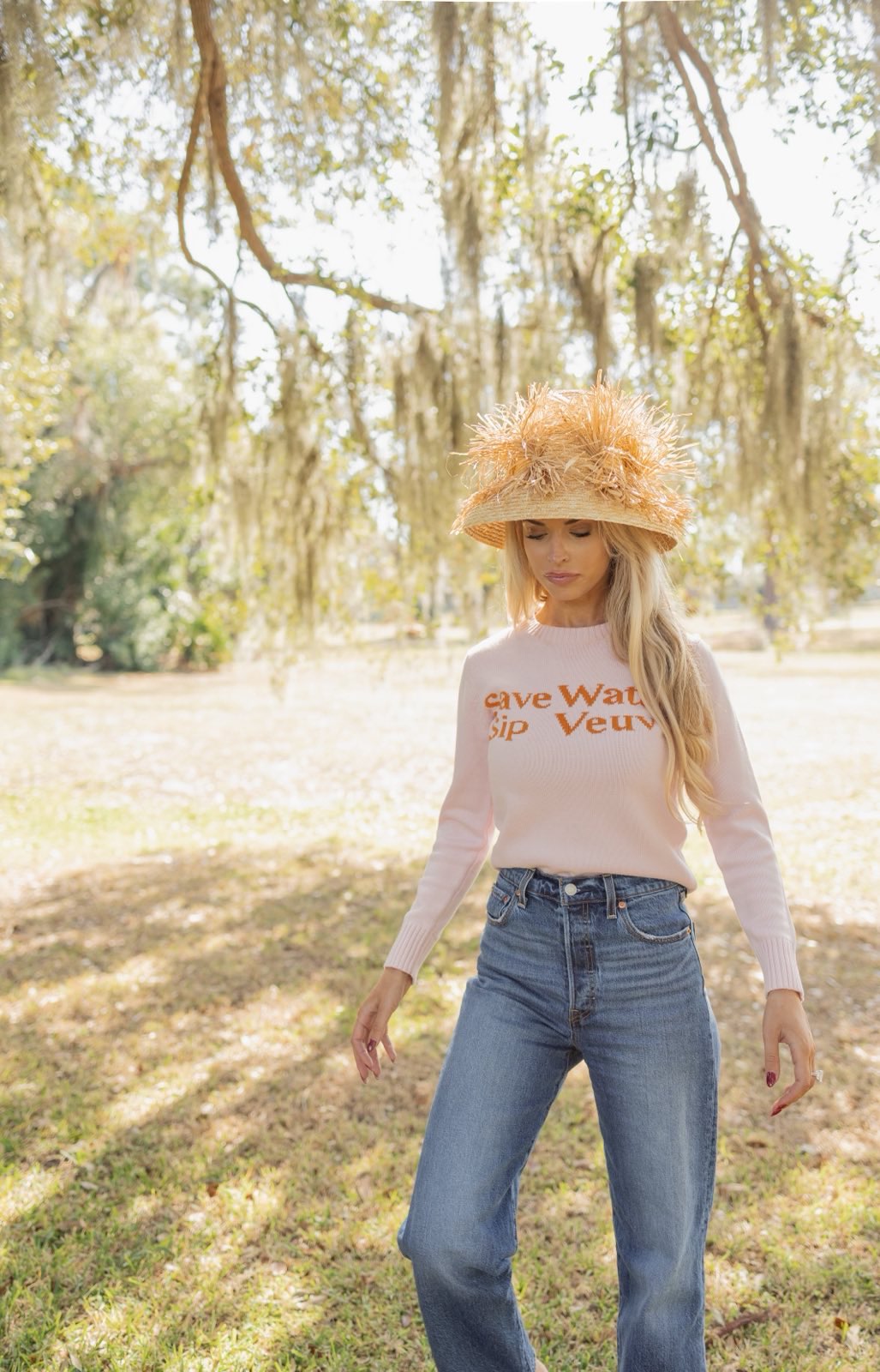 Woman wearing a light pink sweater with text and blue jeans standing in a park.